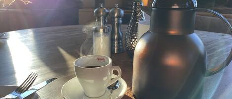 Una mesa con una taza de espresso, una termo, saleros y pimenteros, y una cuchara.