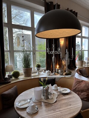 A dining area with a round table set for two, a large black pendant light, and a view of the outdoors through large windows. - Hotel an der Burg (Wettenberg)