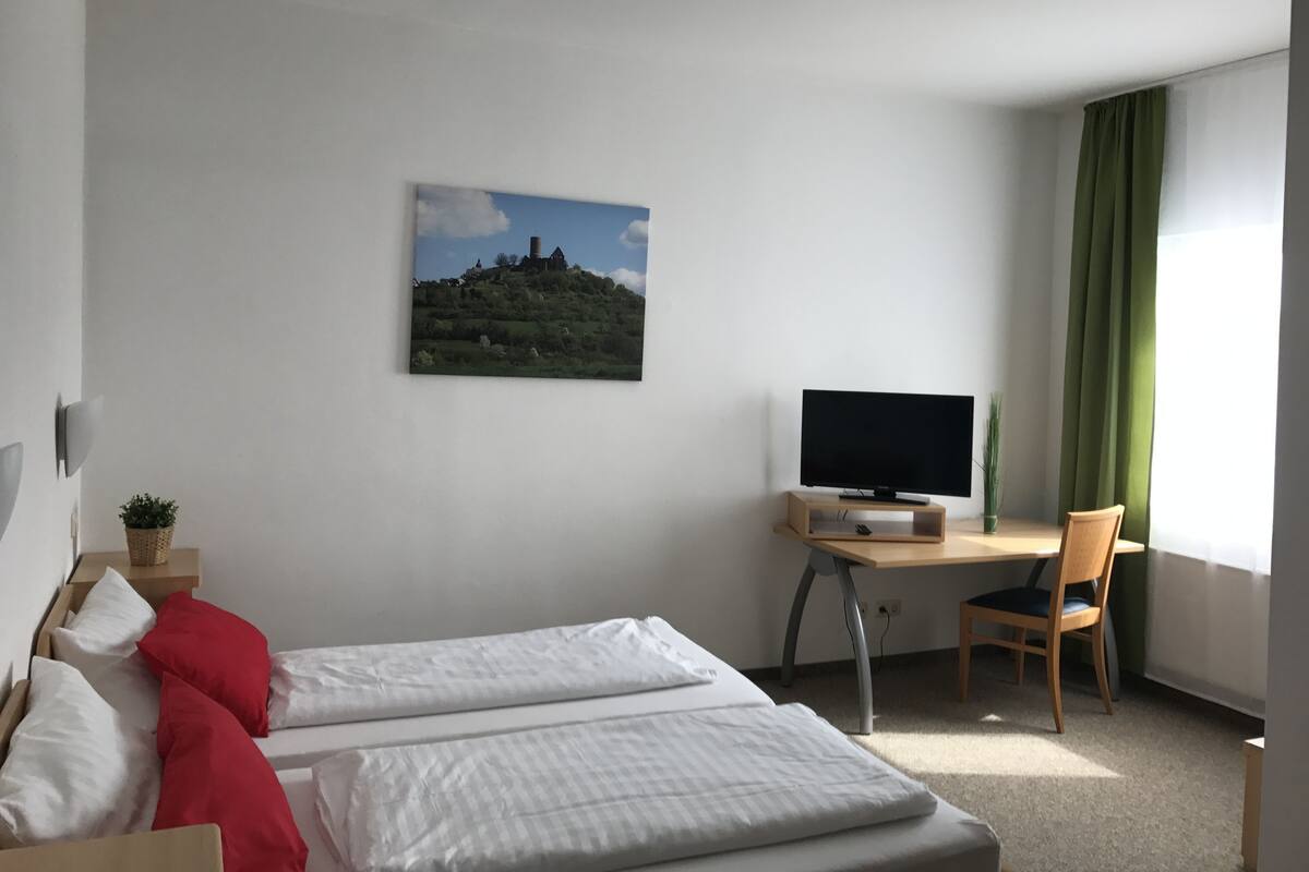 Ein Hotelzimmer mit Bett, Schreibtisch mit Fernseher, Stuhl, einem Fenster mit Vorhängen und einem Bild an der Wand.