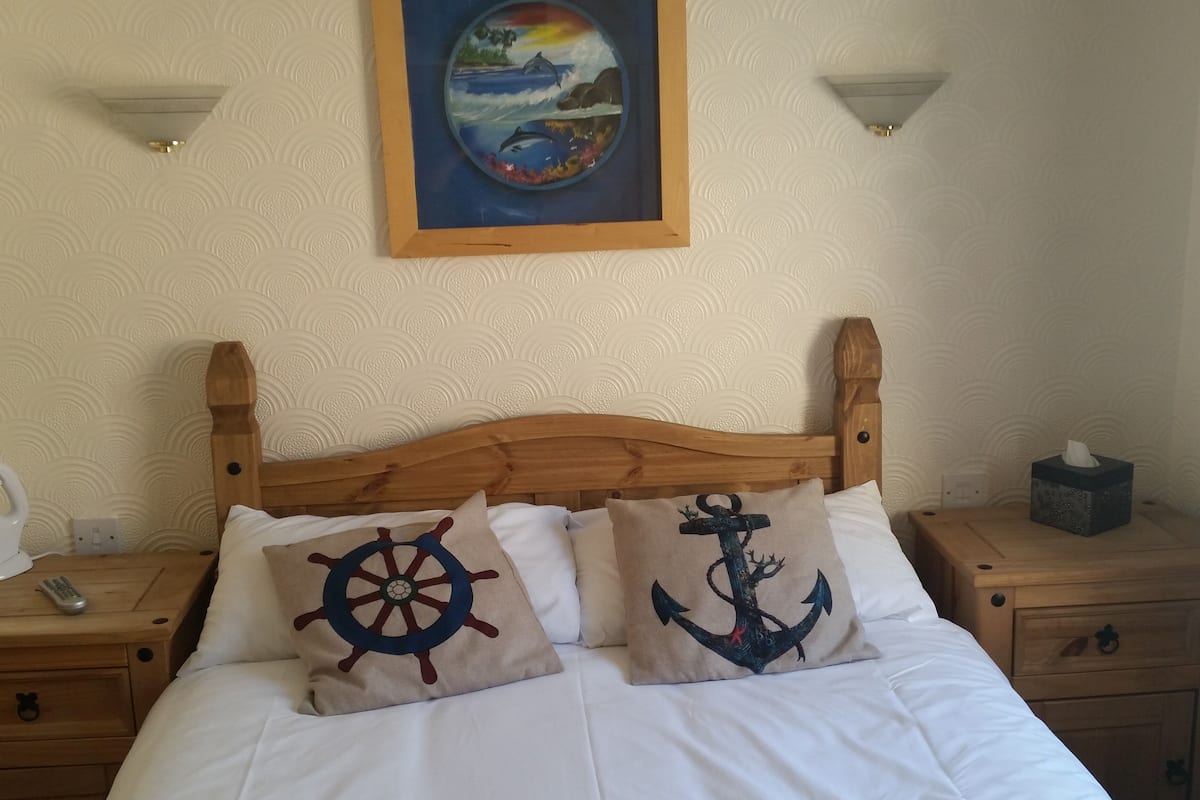 Una cama con dos almohadones decorativos, un cabecero de madera, una mesita de noche con un control remoto y una mesilla con una caja de pañuelos.