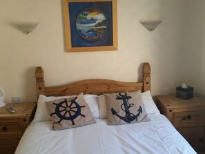 A bed with two decorative pillows, a wooden headboard, a nightstand with a remote control, and a bedside table with a tissue box. - Le Maitre (Southport)