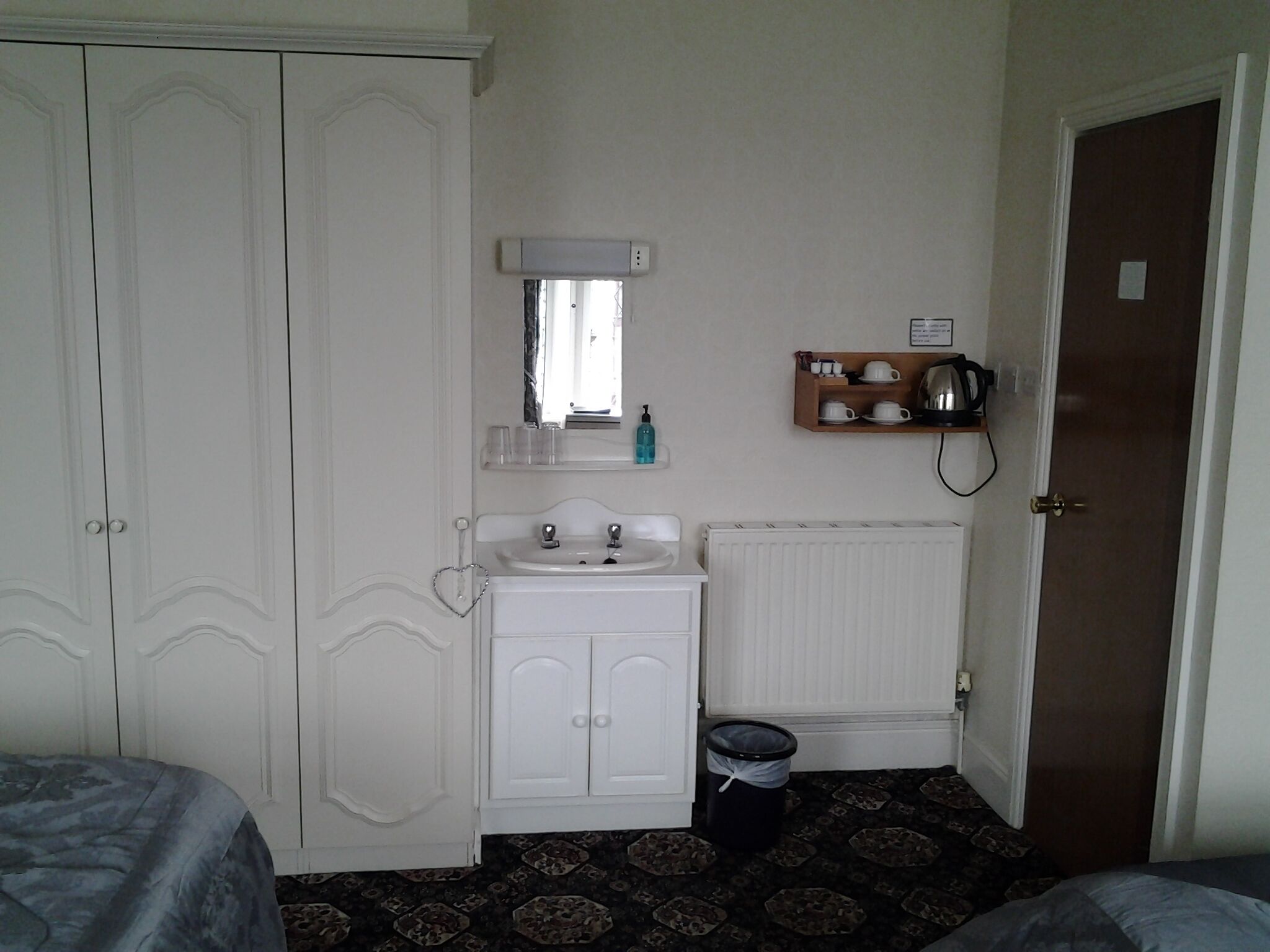 A small, neatly arranged bedroom with a bed, a bedside table, a mirror, a radiator, and a shelf with cups and a kettle.