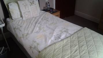 A neatly made bed with floral bedding and a quilted cover. There is a nightstand with a water bottle and a glass beside the bed.
