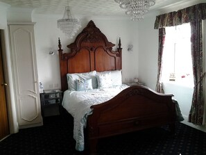 A bedroom with a wooden bed, a nightstand, a chandelier, and a window with curtains. - Le Maitre (Southport)