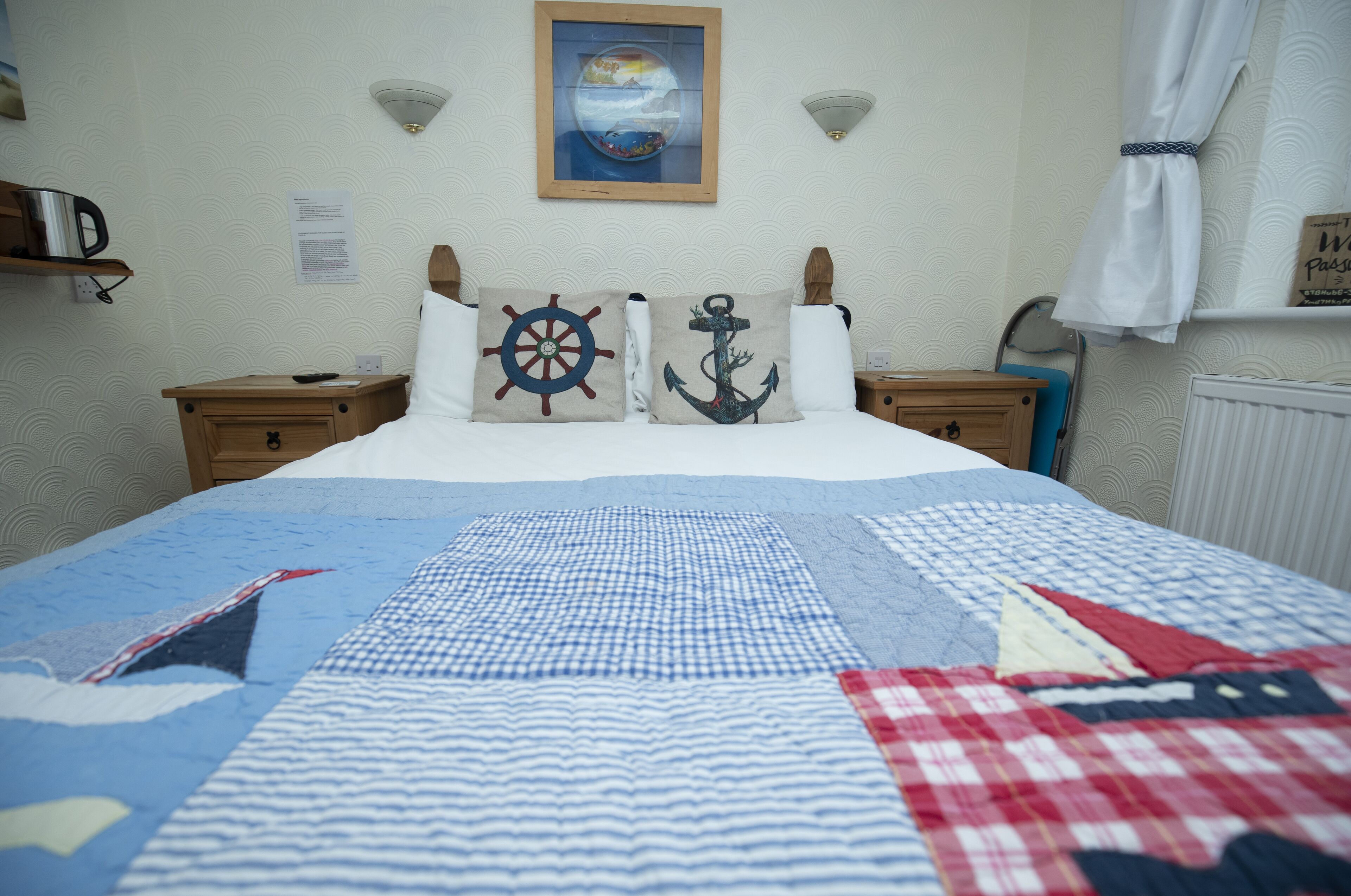 Una cama bien hecha con una colcha azul y blanca a cuadros, dos almohadas con motivos náuticos, un cuadro con un globo terráqueo en la pared y una pequeña estantería con un hervidor.