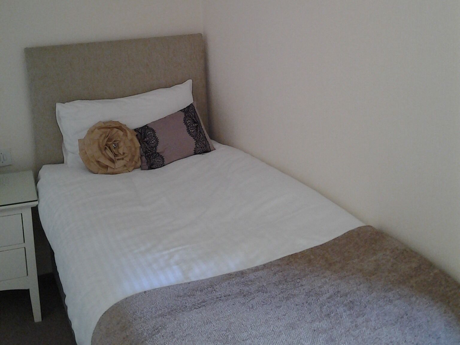 A neatly made bed with a white and brown blanket, a beige headboard, and a decorative pillow.