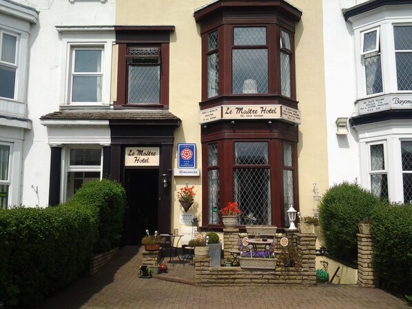 A corner building with a sign for Le Maitre Hotel, featuring a traditional design with a bay window and a small outdoor seating area with potted plants. - Le Maitre (Southport)