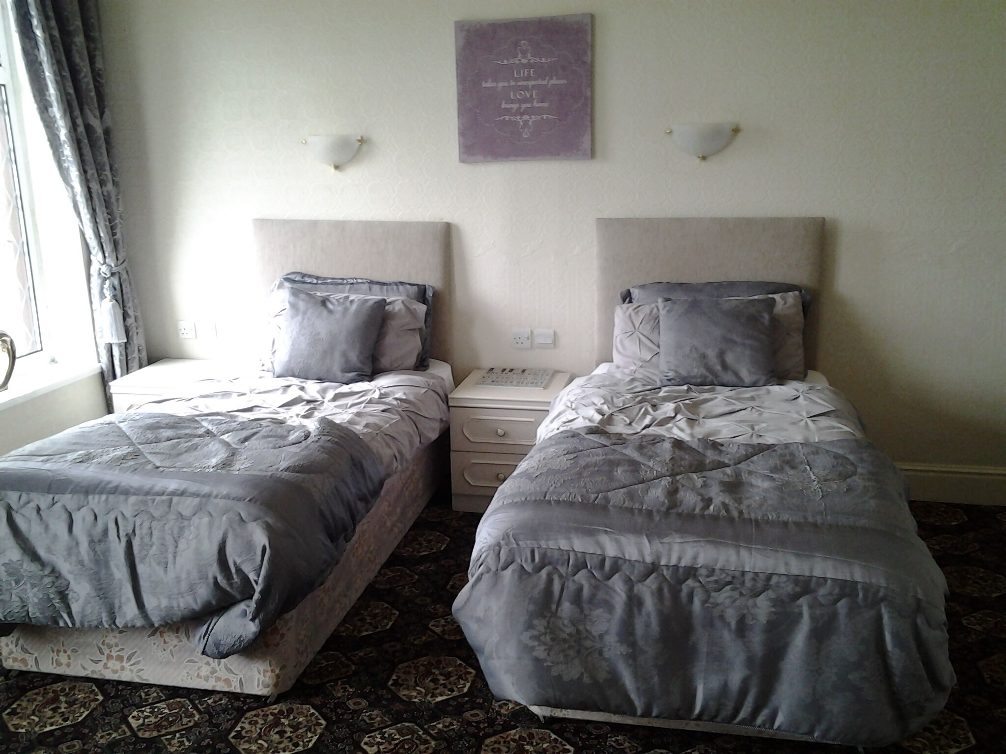 Una habitación con dos camas individuales, una mesita de noche con lámpara, una luz de pared y una placa decorativa.