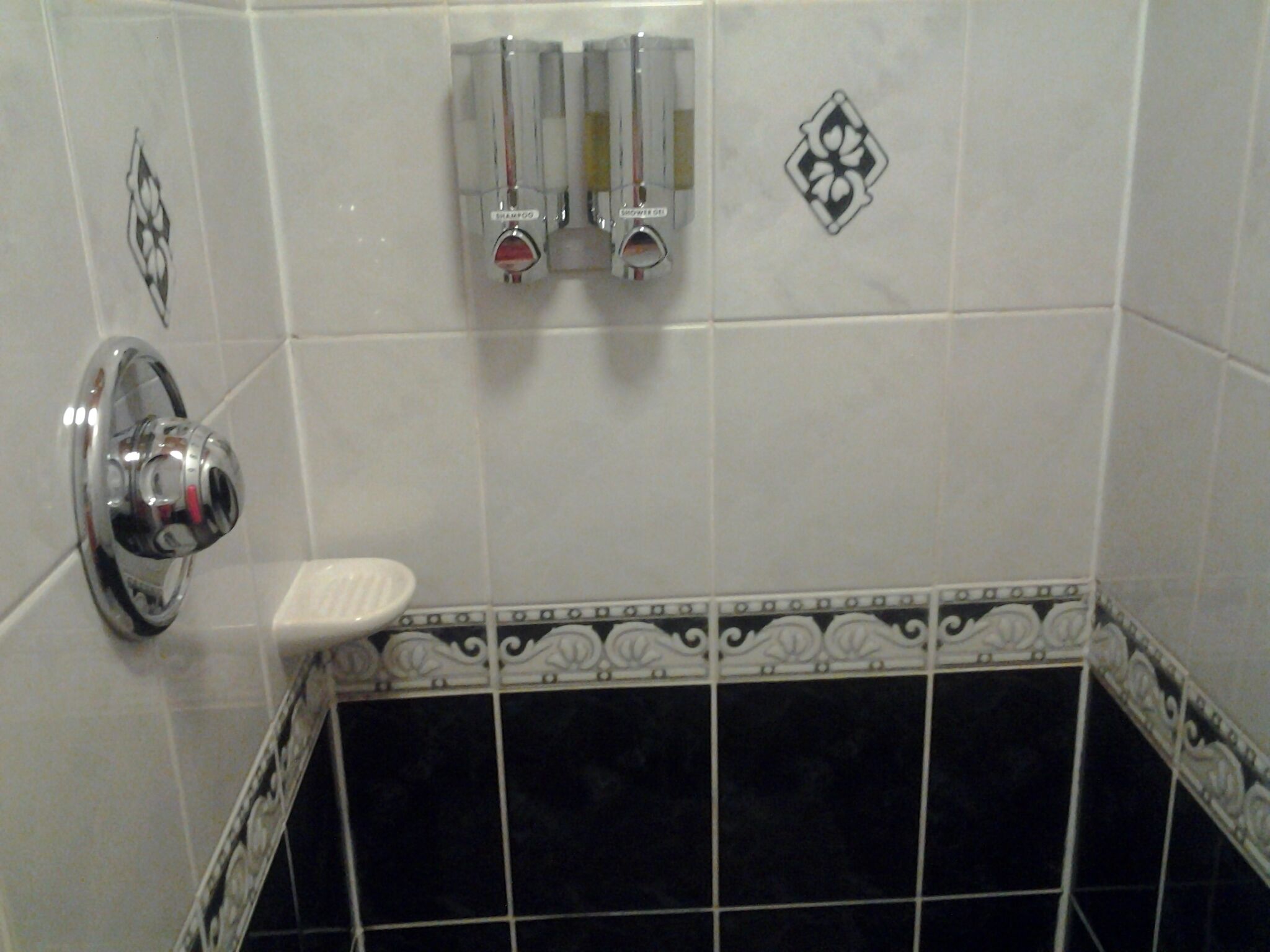 A bathroom with a white tiled wall, a black tiled floor, a chrome faucet, and a soap dispenser.