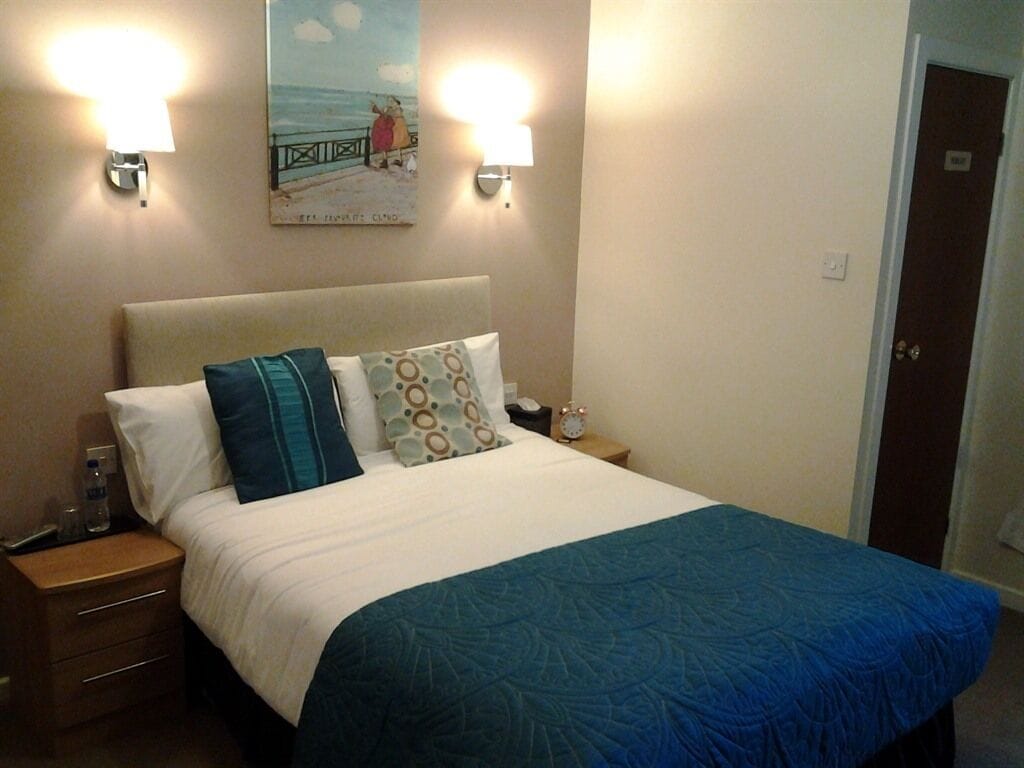A hotel room with a bed, two bedside lamps, a nightstand, a painting on the wall, and a door.