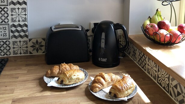 A kitchen counter with a toaster, kettle, croissants, and a fruit bowl.