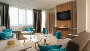 A modern hotel room with a sofa, armchairs, a coffee table, a flat-screen TV mounted on a wooden panel, and a suitcase on the floor. - Oasis del Sol (Tsarevo)