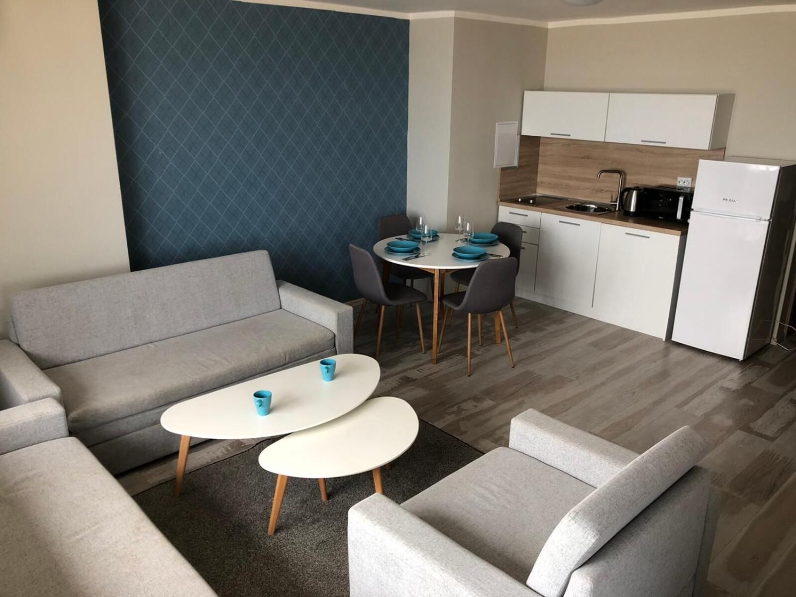 Una sala moderna con un sofá gris, dos mesas de centro blancas y sillones grises. Una mesa de comedor con platos y vasos azules está colocada frente a una pared azul. Se ve una cocina con armarios blancos y un refrigerador negro al fondo.