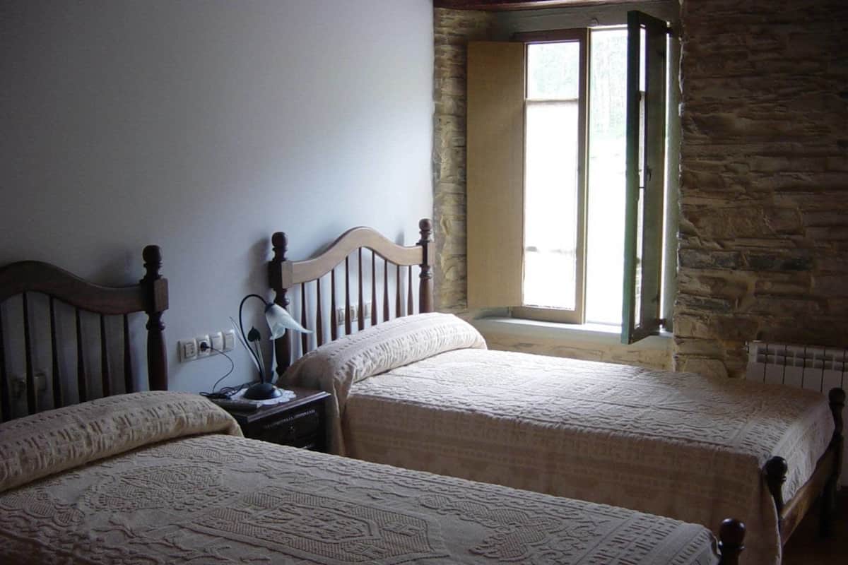 A room with two beds, a stone wall, a window, and a nightstand with a lamp.