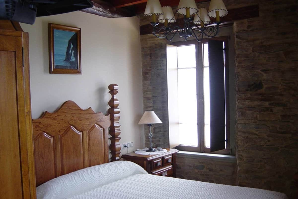 A bedroom with a wooden headboard, a bed with a white quilt, a nightstand with a lamp, a stone wall, and a window with curtains.