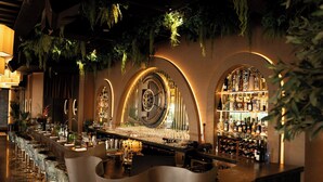 Una zona de bar con una barra alargada, diversas opciones de asientos y una estantería para vinos.
