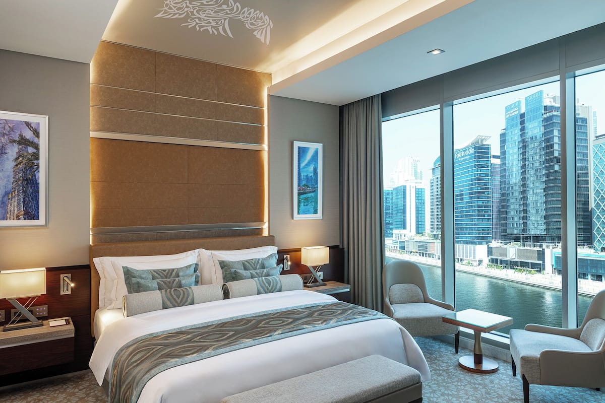 Ein modernes Hotelzimmer mit einem großen Bett, einer Sitzecke mit zwei Sesseln, Blick auf die Stadt durch große Fenster und einer dekorativen Wandverkleidung.
