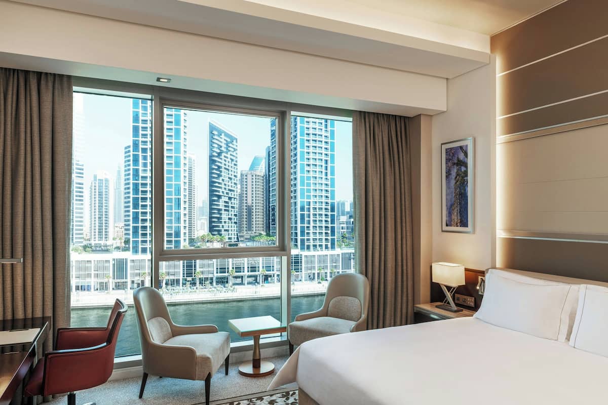 Ein Hotelzimmer mit einem Bett, zwei Sesseln, einem kleinen Tisch, einem Nachttisch und einem großen Fenster mit Blick auf die Stadt.