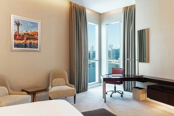 A hotel room with a bed, two armchairs, a desk with a chair, a large window with curtains, a framed cityscape picture, and a minibar.