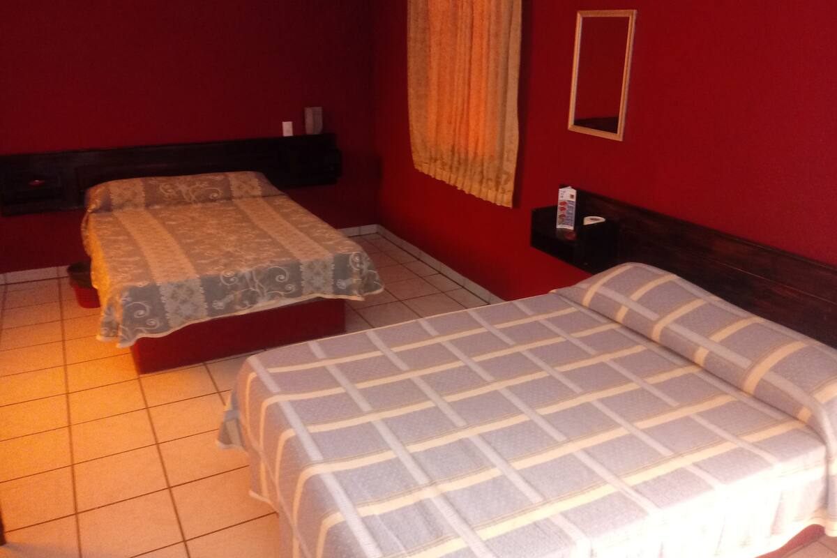 Una habitación con dos camas, una pared roja, un cabecero de madera, una mesita de noche y un espejo.