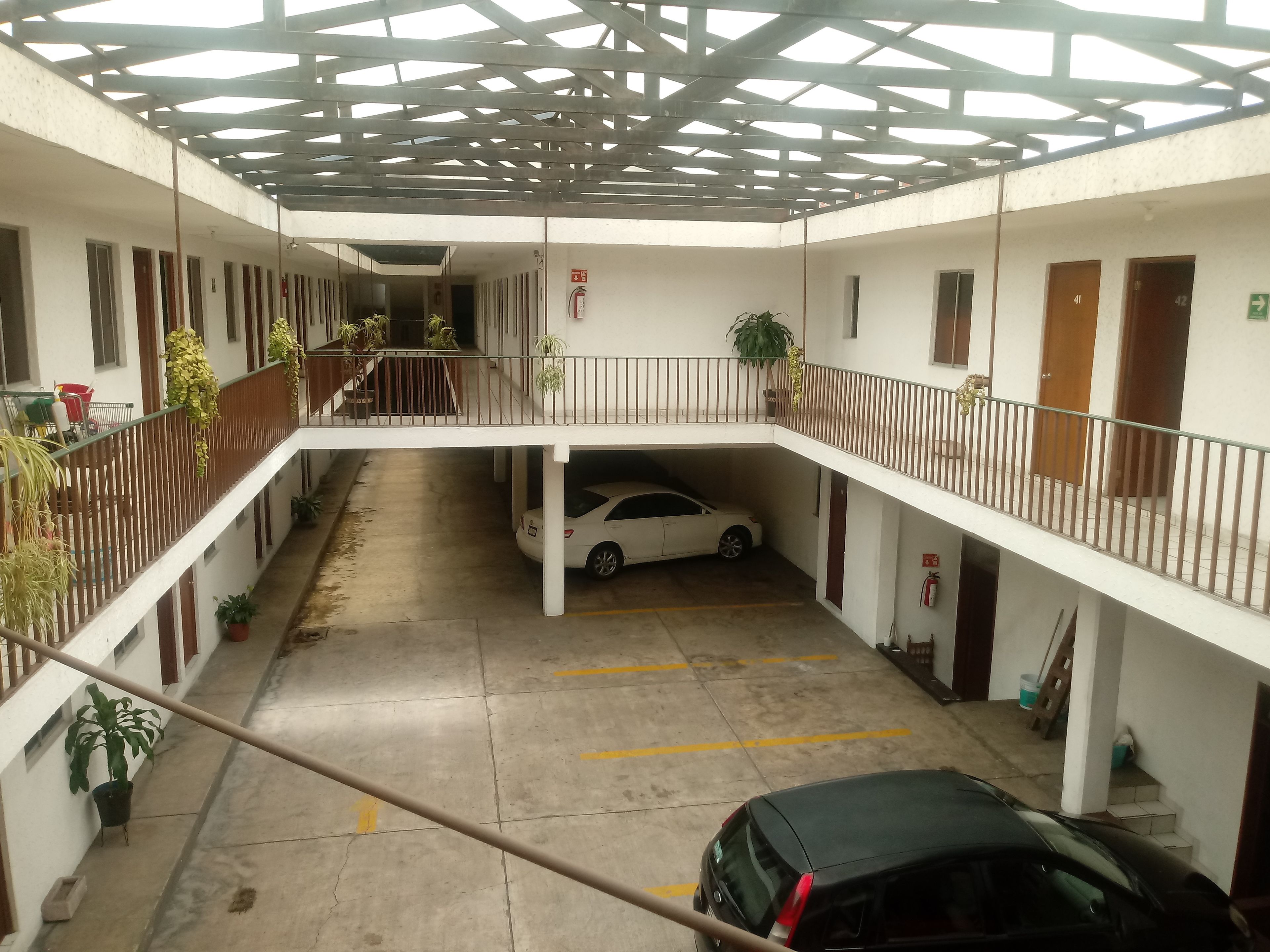 Un aparcamiento de varios niveles con un coche blanco y un coche negro estacionados. Hay plantas en macetas a lo largo de las barandillas y un extintor de incendios montado en la pared.