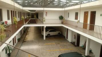 Un aparcamiento de varios niveles con un coche blanco y un coche negro estacionados. Hay plantas en macetas a lo largo de las barandillas y un extintor de incendios montado en la pared.