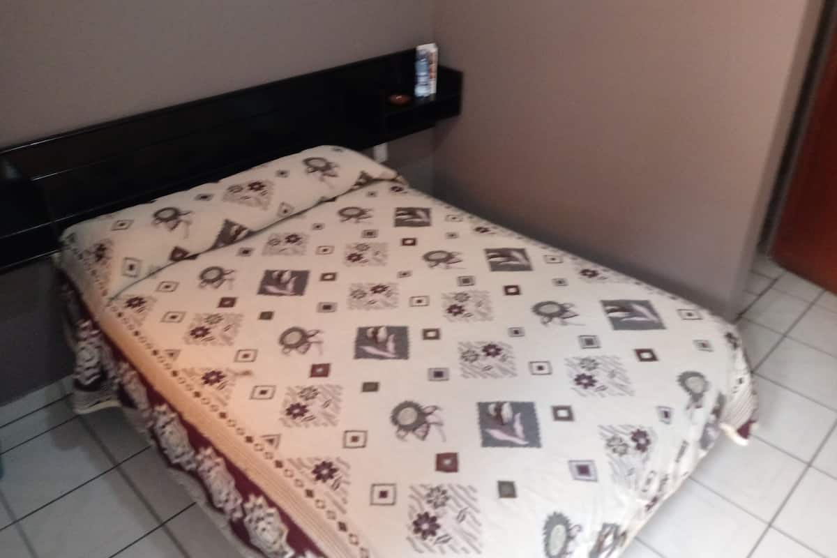 Una cama pequeña y bien hecha con una colcha estampada en una habitación con suelo de baldosas y cabecero oscuro.