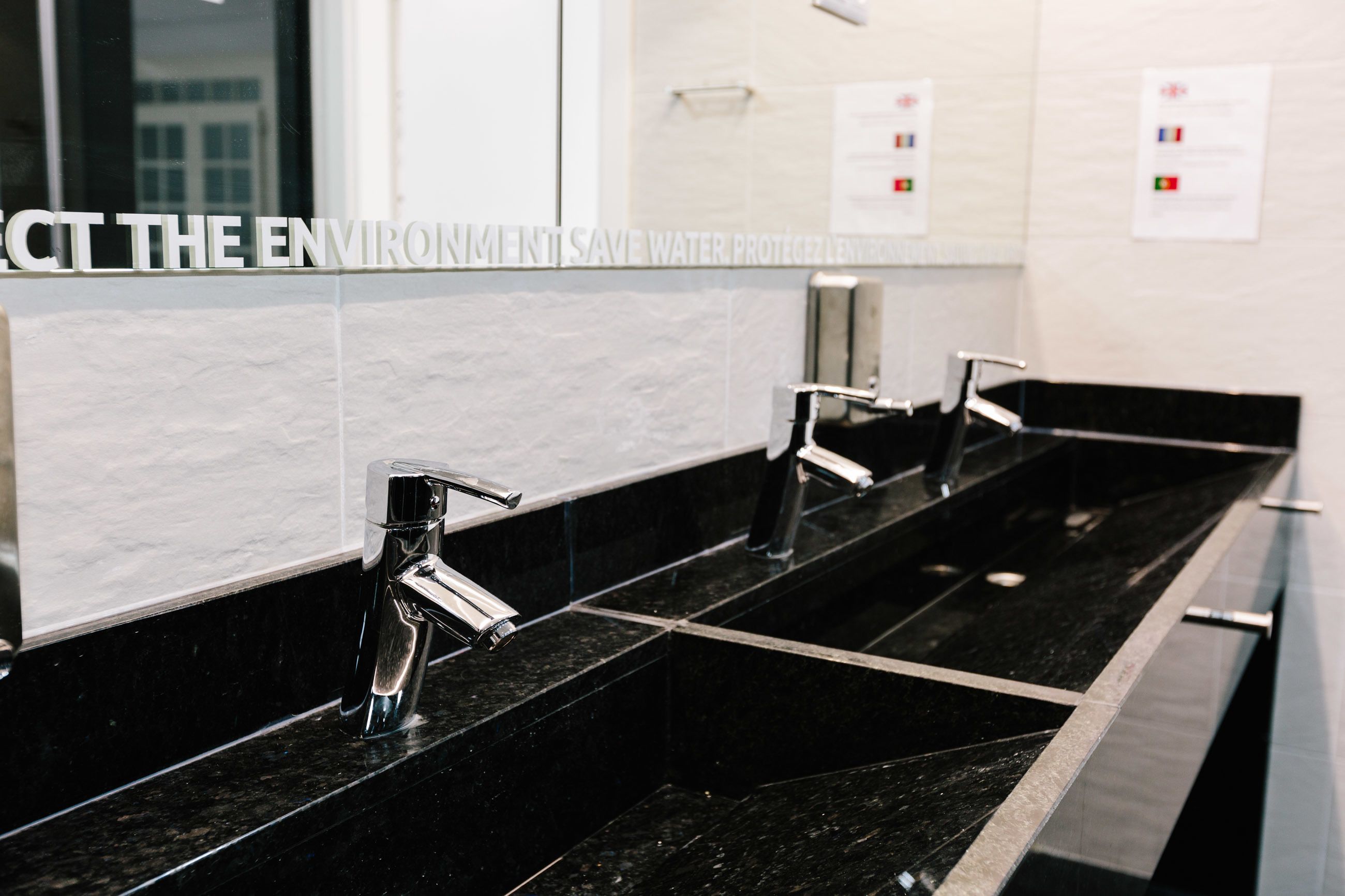 A modern bathroom with three sinks, black marble countertops, and a mirror with a message about water conservation.