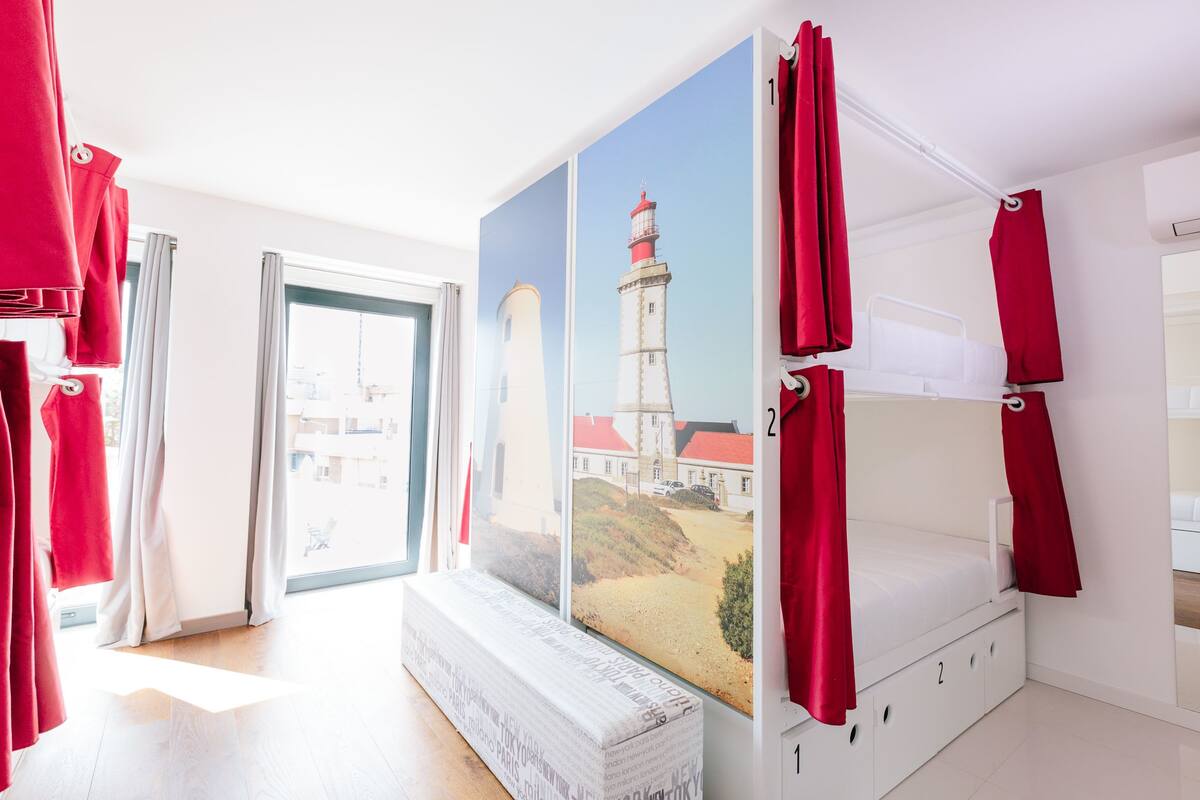A room with a bunk bed, a large window with a view of a lighthouse, and red curtains.