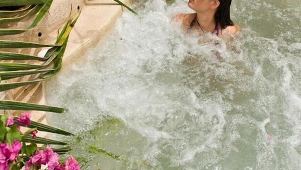 A person in a hot tub surrounded by pink flowers and green plants.