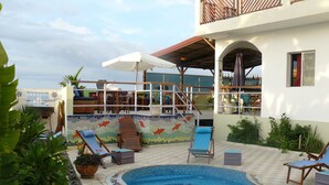 Una piscina con mosaicos, rodeada de sillas de descanso y sombrillas, con vista a un edificio y un balcón al fondo.