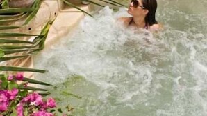 Una persona en un jacuzzi con chorros de agua, rodeada de plantas verdes y flores rosadas.