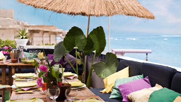 Una mesa de comedor servida con platos, vasos y un jarrón de flores, con vista al mar.