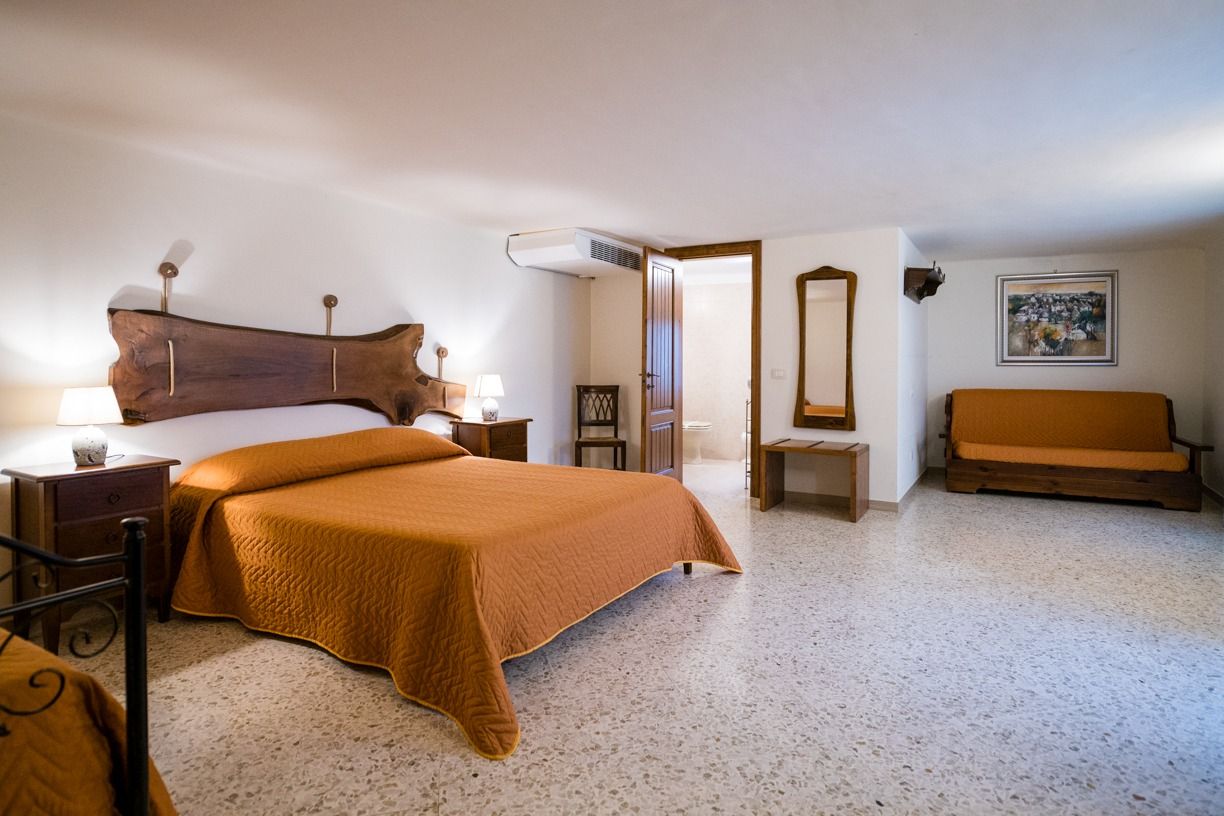 Un dormitorio con una cama grande, mesitas de noche, un banco de madera, un espejo y una puerta que da a otra habitación.