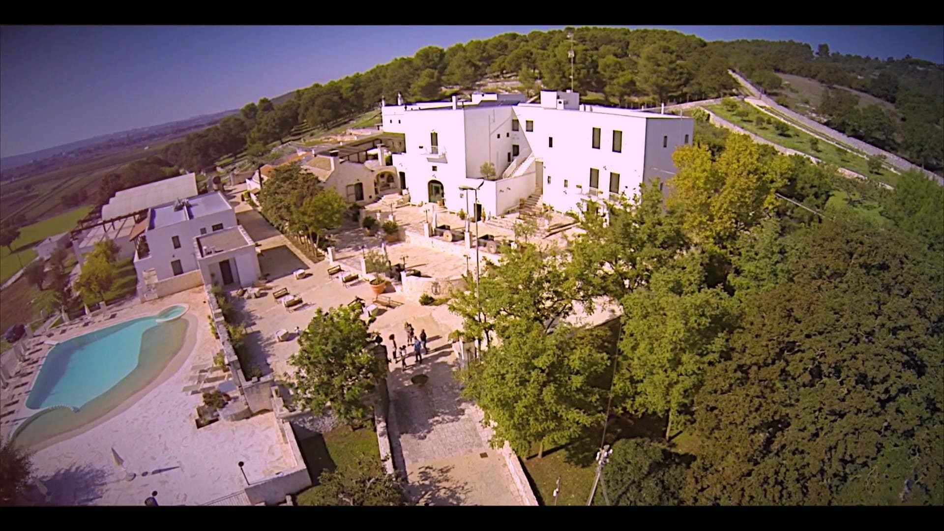 A large white building with multiple floors, surrounded by lush greenery and a swimming pool.