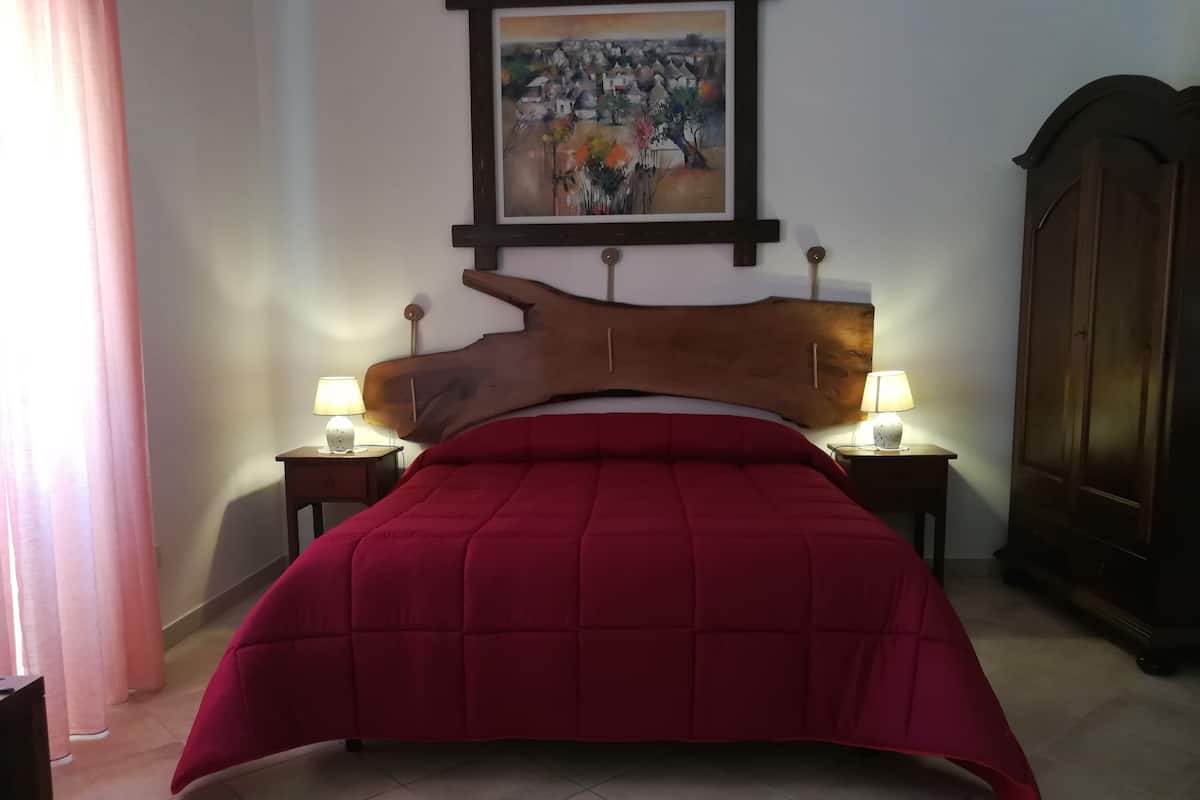 Ein Schlafzimmer mit hohem, hölzernen Kopfteil, einer roten Bettdecke, zwei Nachttischlampen, einem Bild an der Wand und einem Kleiderschrank.