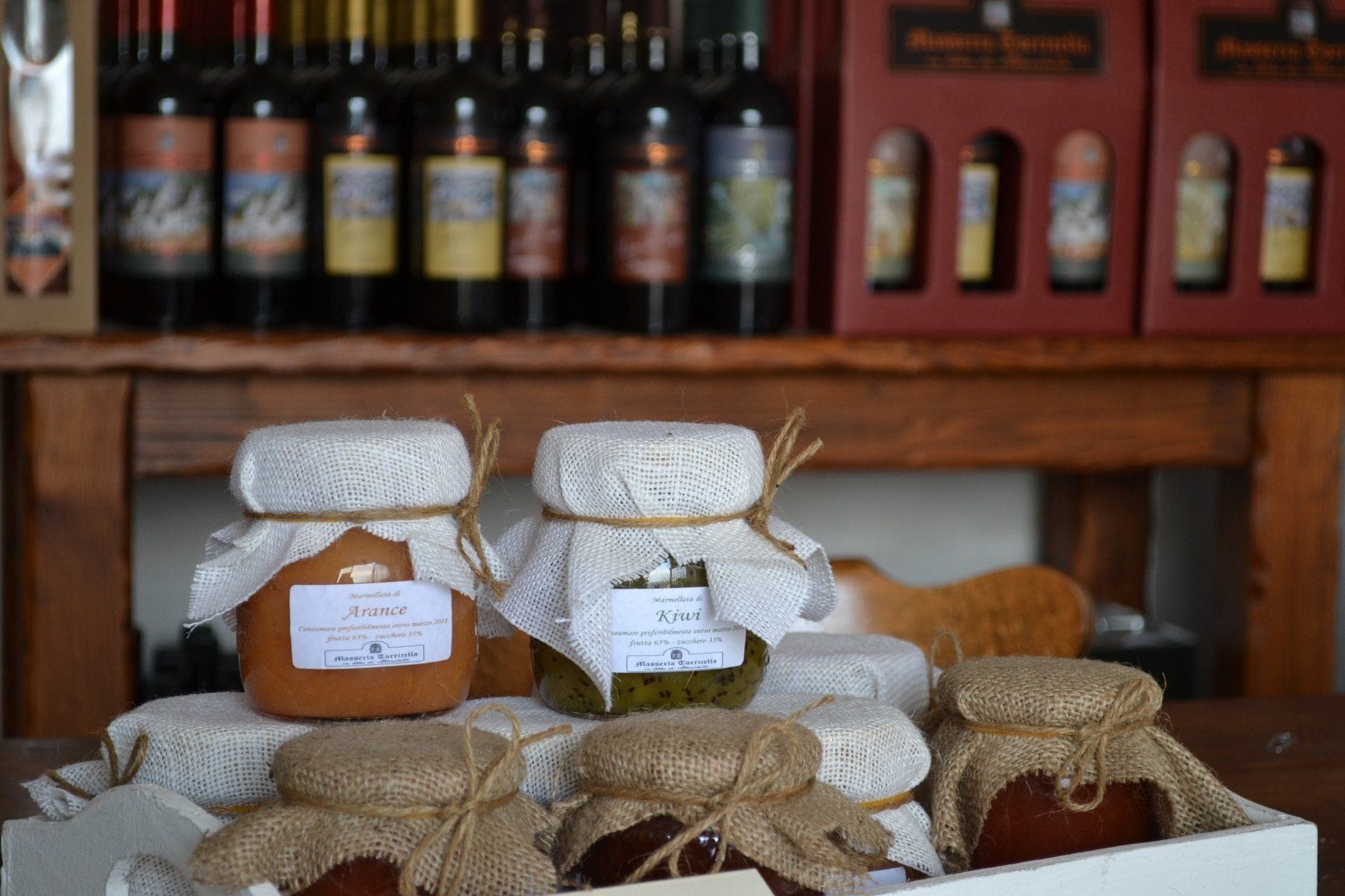 Jars of homemade preserves on a table, with a backdrop of wine bottles and a wooden shelf.