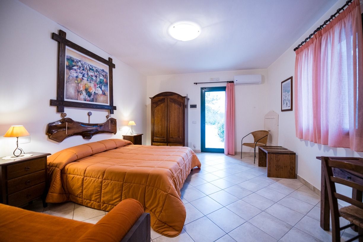 A bedroom with a large bed, a wooden wardrobe, a desk, and a chair. There is a door leading to an outdoor area.