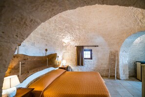 A rustic bedroom with a stone wall, a wooden bed, a nightstand, a lamp, and a window.