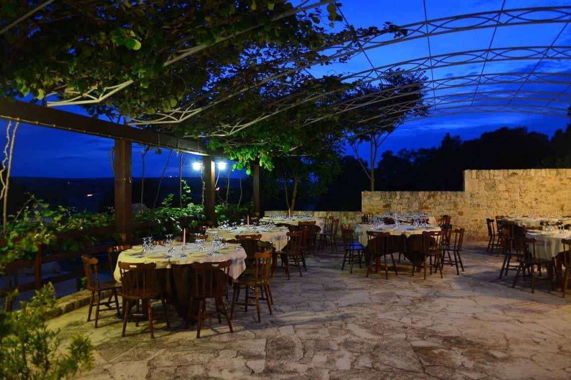 An outdoor dining area with tables set for a meal, under a pergola with string lights, overlooking a body of water.