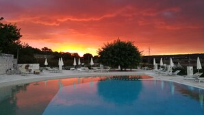 Une piscine entourée d’arbres, avec des chaises longues et des parasols, le tout baigné par la lumière d’un coucher de soleil.
