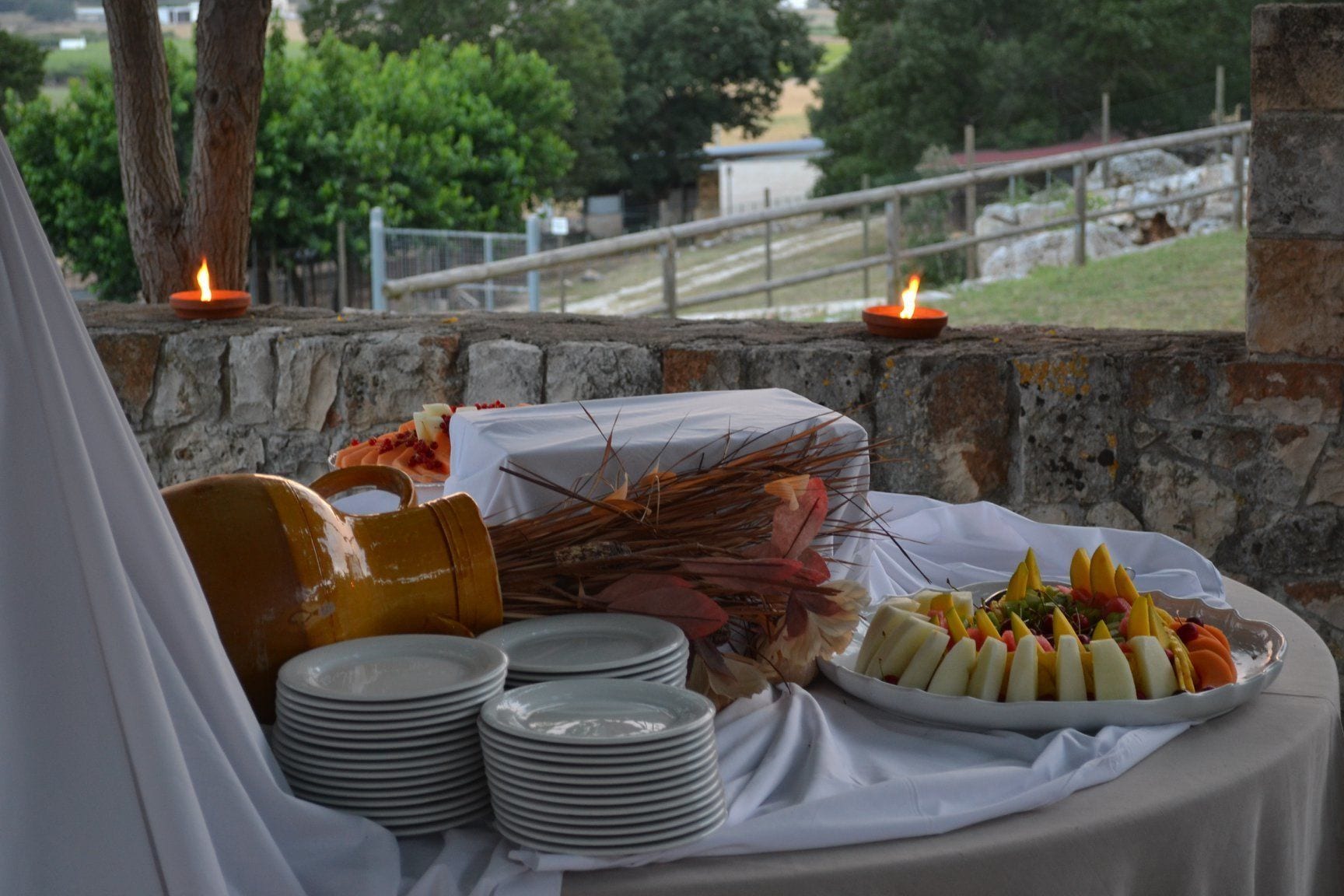 Un montaje para comer al aire libre con un muro de piedra de fondo, una mesa cubierta con un mantel blanco, platos, una jarrón y una bandeja de frutas.