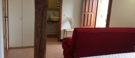 Une chambre à coucher avec un lit, un pilier en bois, une chaise rouge, une porte en bois et une vue sur une salle de bain avec un lavabo et un miroir.