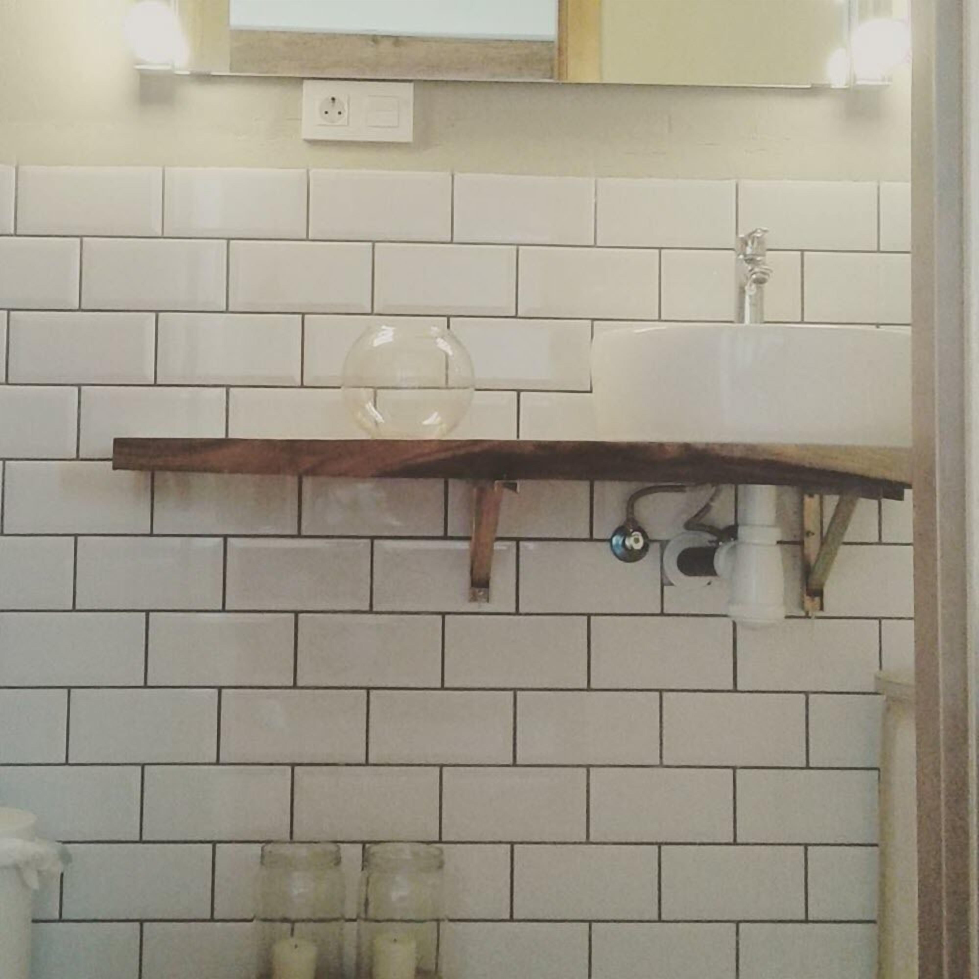A bathroom with white tiled walls, a wooden shelf above the sink, and a mirror above the shelf.