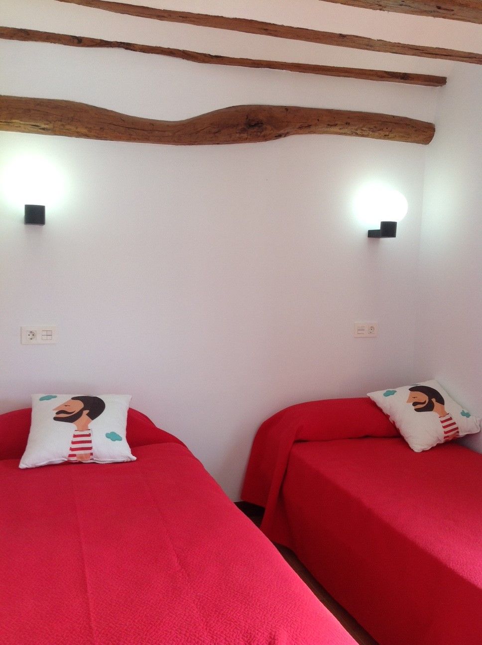 Een kamer met twee bedden, elk met een rode spreideken en een kussen met een tekenfilmfiguur. De kamer heeft zichtbare houten balken aan het plafond en twee wandlampen.