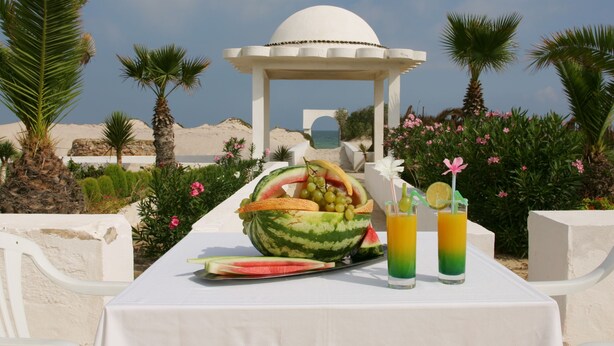 A table with a watermelon bowl, fruit slices, and two drinks with fruit garnishes, set against a backdrop of palm trees and a dome structure. - Hôtel El Andalous (Soliman)