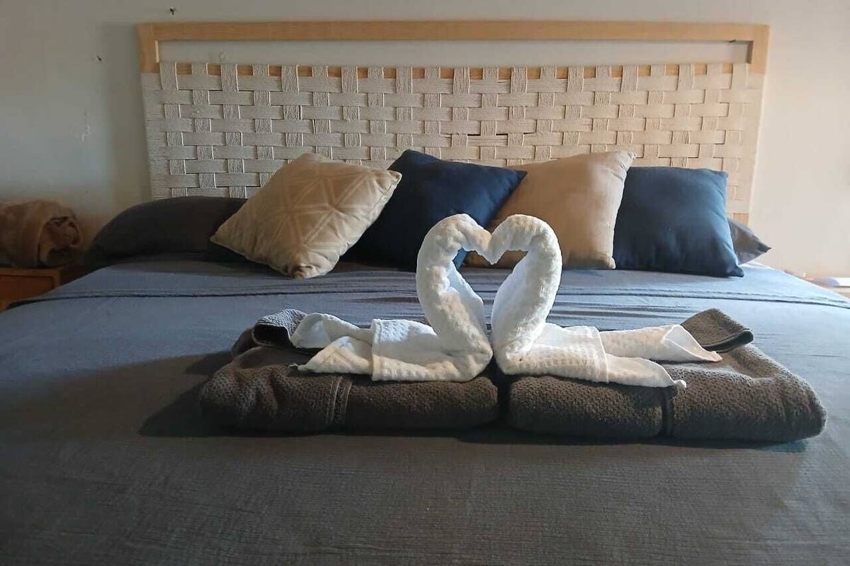 A neatly made bed with a woven headboard, two pillows, and a heart-shaped towel arrangement.