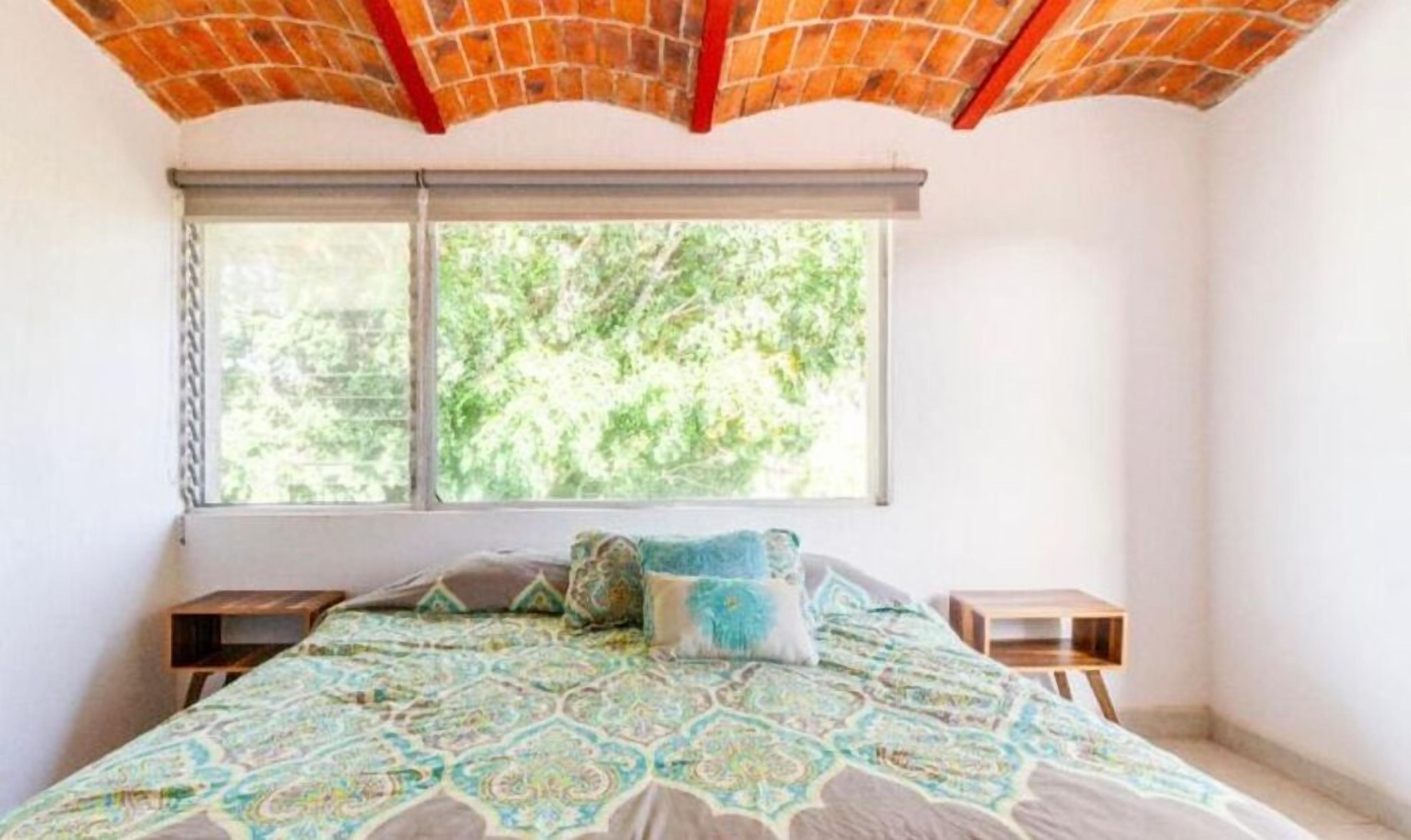 Un dormitorio con colcha estampada, ventana con vistas a la vegetación y techo de ladrillo.