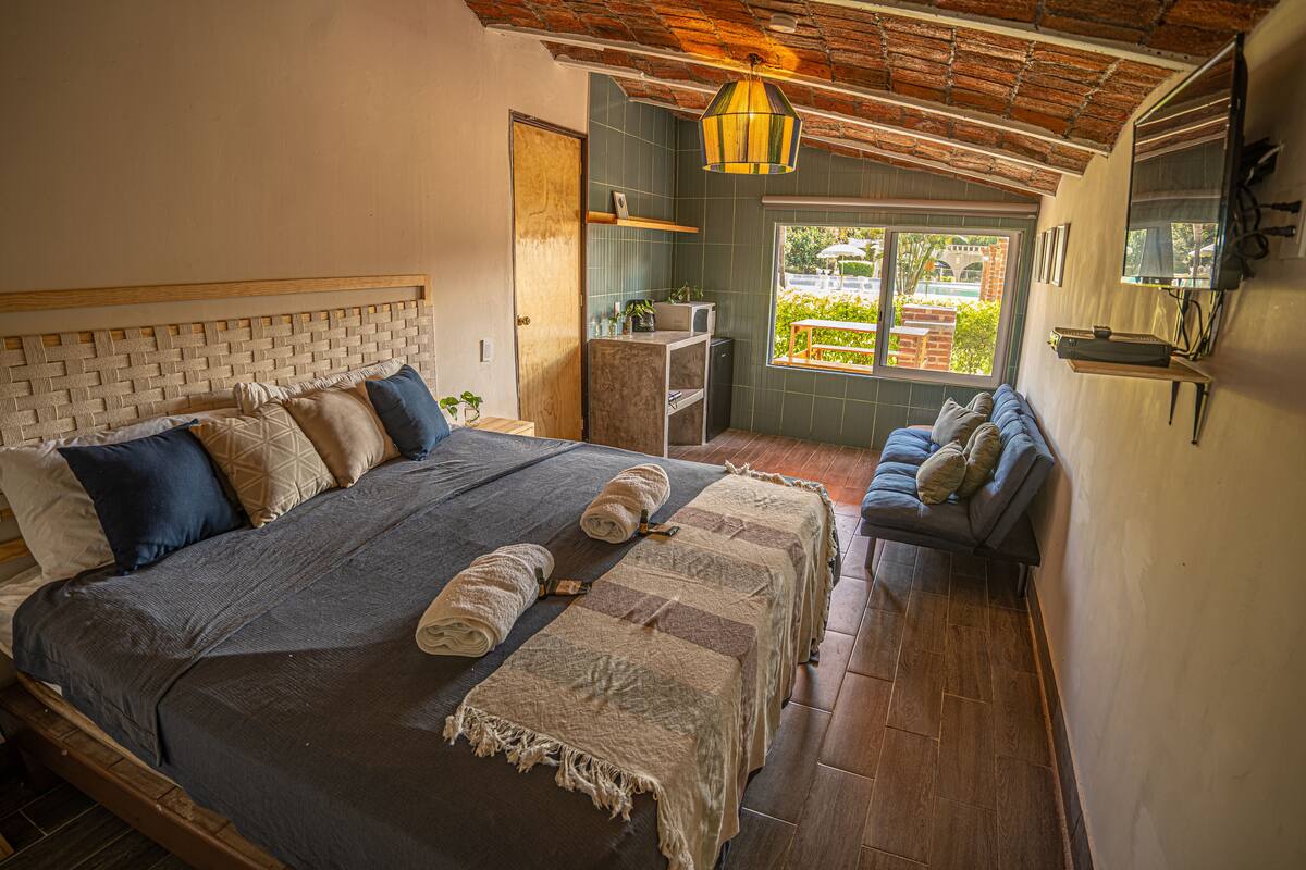 A bedroom with a large bed, a bench, a desk, and a window with a view of the outside.