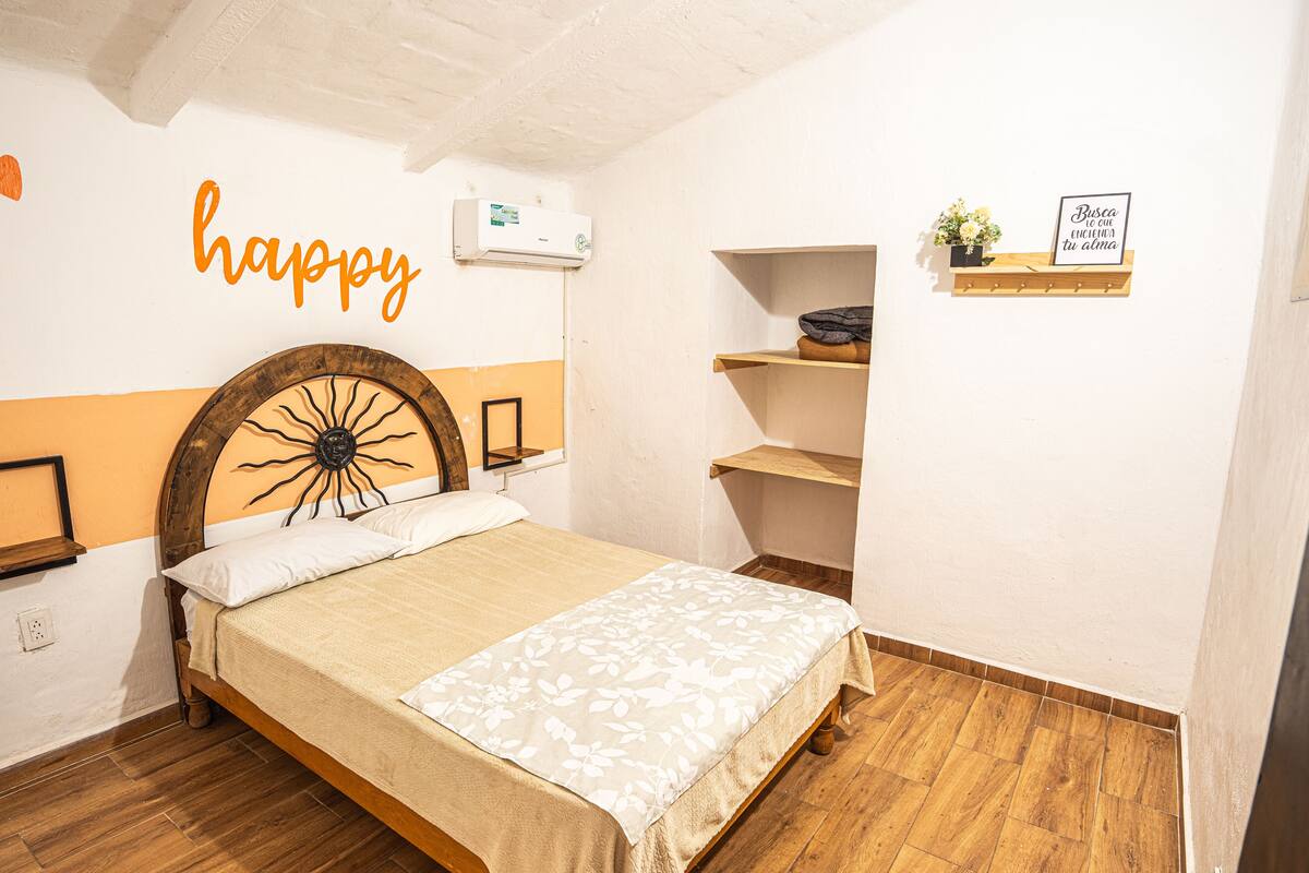 A bedroom with a bed, a wall-mounted air conditioner, a shelf with a potted plant, and a framed sign.